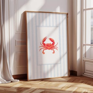 May include: A framed print featuring a red crab illustration. The artwork is set against a backdrop of blue and white striped paper, framed in gold. The crab is centered on a white rectangle with a blue border.