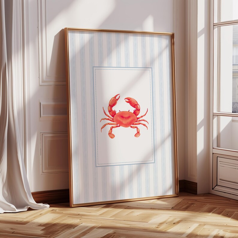 May include: A framed print featuring a red crab illustration. The artwork is set against a backdrop of blue and white striped paper, framed in gold. The crab is centered on a white rectangle with a blue border.