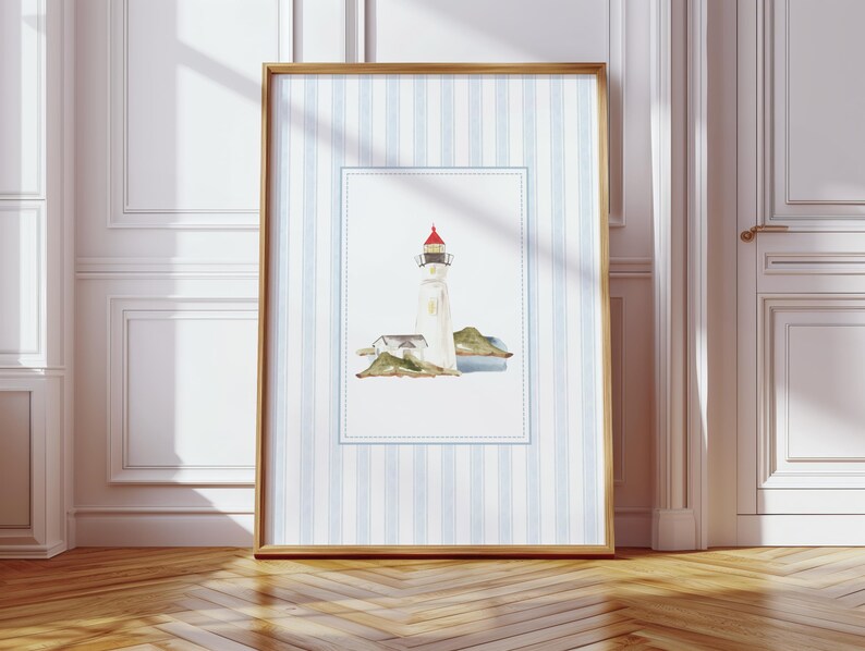 May include: Framed watercolor print of a lighthouse with a red roof, set on a small island. The artwork is framed in a wooden frame and set against a blue and white striped background. The frame is displayed on a wooden floor.