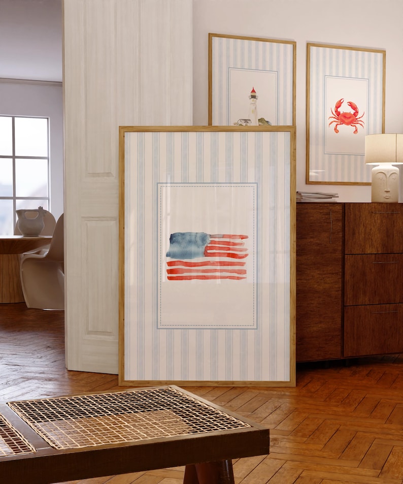 May include: Watercolor artwork of the American flag in a wooden frame. The flag features red and blue watercolor strokes. Two additional framed artworks with nautical themes are visible in the background. The frames are displayed against a blue and white striped background.