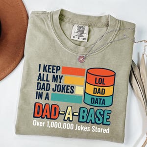 May include: A light olive green t-shirt featuring a graphic with the text I KEEP ALL MY DAD JOKES IN A DAD-A-BASE Over 1,000,000 Jokes Stored. The design includes a stylized database icon with orange, yellow, and teal sections labeled LOL, DAD, DATA.