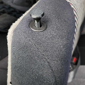 May include: A close-up of a grey car seat with a black plastic button. The button is round and has a textured surface. The seat is made of a soft, grey fabric.