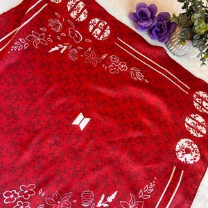 Può includere: Un foulard quadrato rosso con un motivo damascato rosso scuro ripetuto, disegni floreali bianchi a linee e motivi circolari bianchi lungo i bordi. Un logo geometrico bianco composto da due trapezi è centrato sul tessuto, incorniciato da linee bianche.