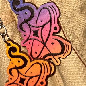 May include: Two wooden pendants with a gradient of colors from red to orange to purple. Each pendant has a black, swirling design with a geometric center and is attached to a silver chain. The pendants are on a tan fabric background.