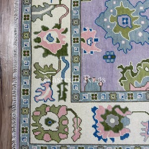 May include: A section of a rectangular rug with a floral pattern. The rug features a light purple field with blue, green, and pink floral designs. The border is white with a blue and green floral pattern. The text "Nasreen Rugs" is visible.