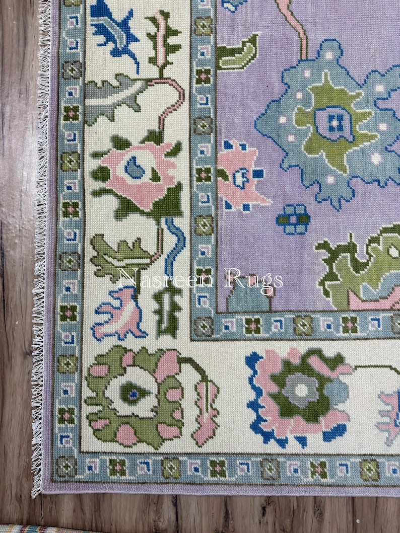 May include: A section of a rectangular rug with a floral pattern. The rug features a light purple field with blue, green, and pink floral designs. The border is white with a blue and green floral pattern. The text "Nasreen Rugs" is visible.