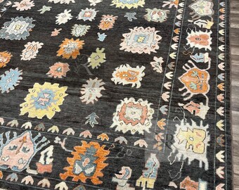 Puede incluir: Una alfombra negra con un patrón geométrico colorido. La alfombra tiene un borde de flecos y presenta una variedad de formas y colores, incluyendo rojo, naranja, amarillo, azul y verde.