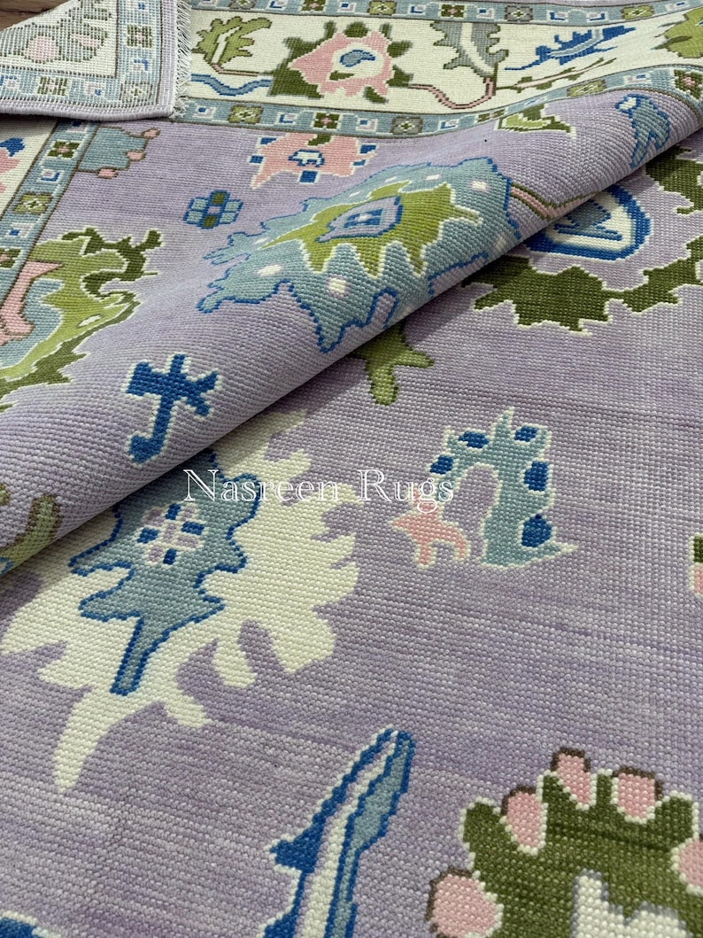 May include: A rolled-up rug with a detailed pattern in lavender, blue, green, and pink. The design includes floral and geometric motifs. The text "Nasreen Rugs" is displayed on the rug.