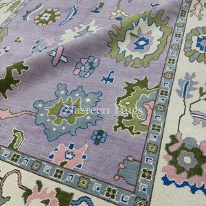 May include: A close-up of a lavender-coloured rug with a floral pattern. The rug features a central field with stylized floral motifs in shades of green, blue, and pink. The border is cream-coloured with a geometric design. The text "Nasreen Rugs" is visible.