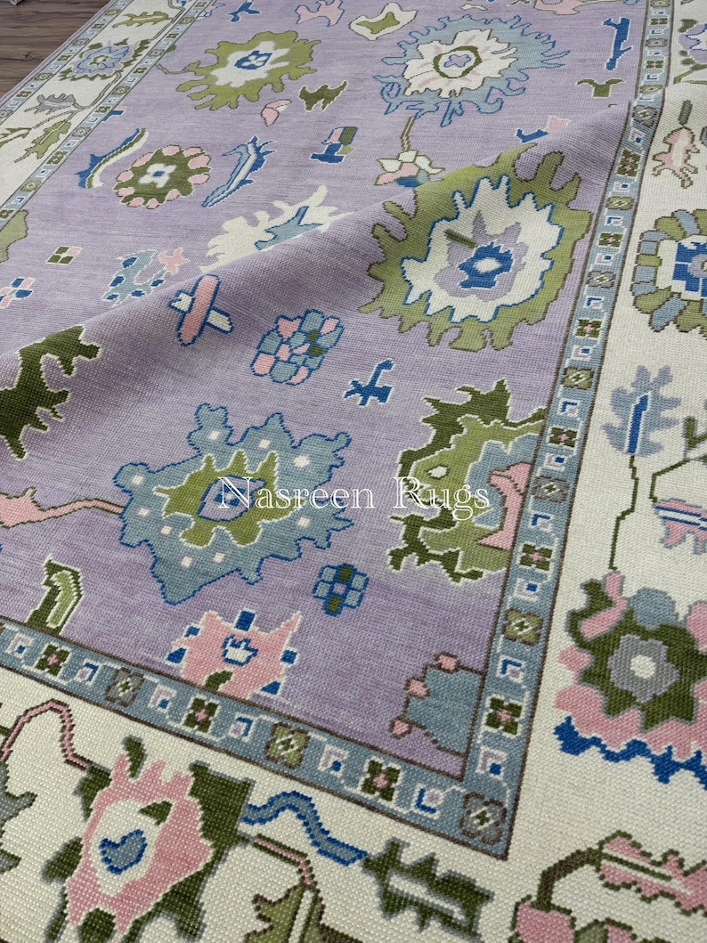 May include: A close-up of a lavender-coloured rug with a floral pattern. The rug features a central field with stylized floral motifs in shades of green, blue, and pink. The border is cream-coloured with a geometric design. The text "Nasreen Rugs" is visible.