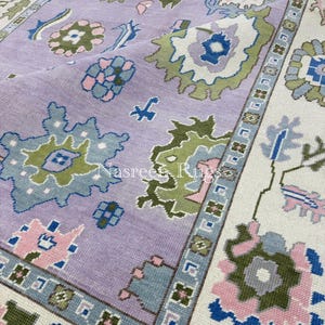 May include: A close-up of a large, rectangular rug with a floral pattern. The rug features a lavender field with green, blue, and pink floral designs. The border is off-white with a repeating geometric pattern. The rug is made of a woven material.