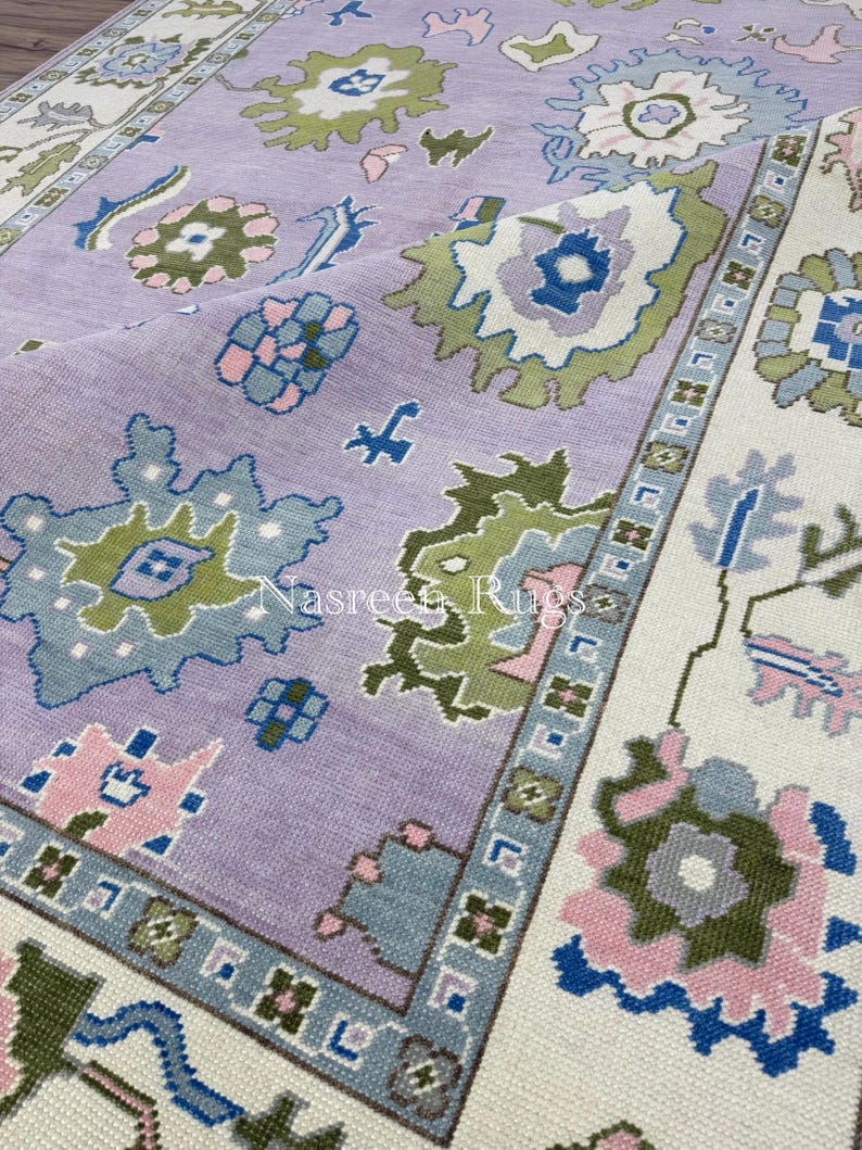 May include: A close-up of a large, rectangular rug with a floral pattern. The rug features a lavender field with green, blue, and pink floral designs. The border is off-white with a repeating geometric pattern. The rug is made of a woven material.