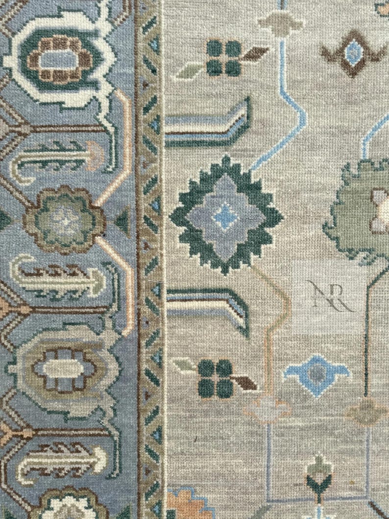 K&ouml;nnte beinhalten: Nahaufnahme eines Teppichs mit hellgrauem Hintergrund und einem geometrischen Muster in Blau, Gr&uuml;n und Braun. Der Teppich hat einen Rand mit einem sich wiederholenden Muster aus Dreiecken und Rauten. Der Teppich ist unten rechts mit "NR" signiert.