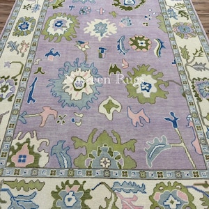 May include: A rectangular rug with a lavender central field and a cream border. The rug displays floral and geometric patterns in green, blue, and pink. The text "Green Rugs" is visible on the rug.