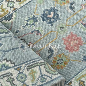 May include: A rolled-up, light blue rug with a detailed floral pattern. The rug features a border with geometric designs in white, green, and gold. The text "Nasreen Rugs" is visible on the rug.