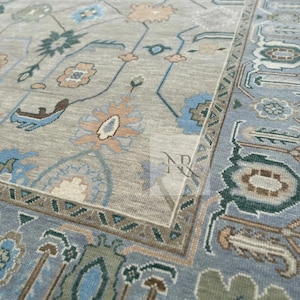 K&ouml;nnte beinhalten: Ein hellblauer Teppich mit einem beigefarbenen Zentrum und komplizierten Blumenmustern in Blaut&ouml;nen, Gr&uuml;n und Beige. Der Teppich hat einen Rand mit einem geometrischen Muster.