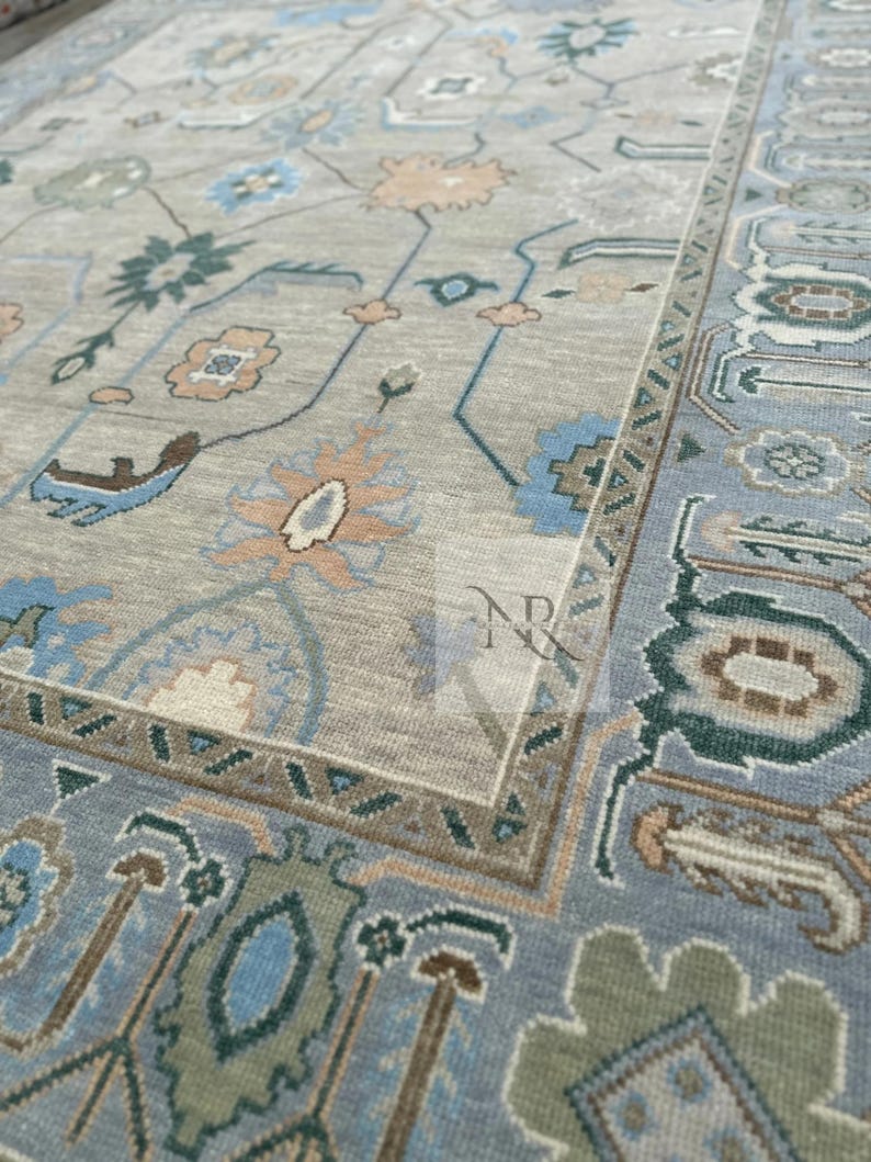 K&ouml;nnte beinhalten: Ein hellblauer Teppich mit einem beigefarbenen Zentrum und komplizierten Blumenmustern in Blaut&ouml;nen, Gr&uuml;n und Beige. Der Teppich hat einen Rand mit einem geometrischen Muster.