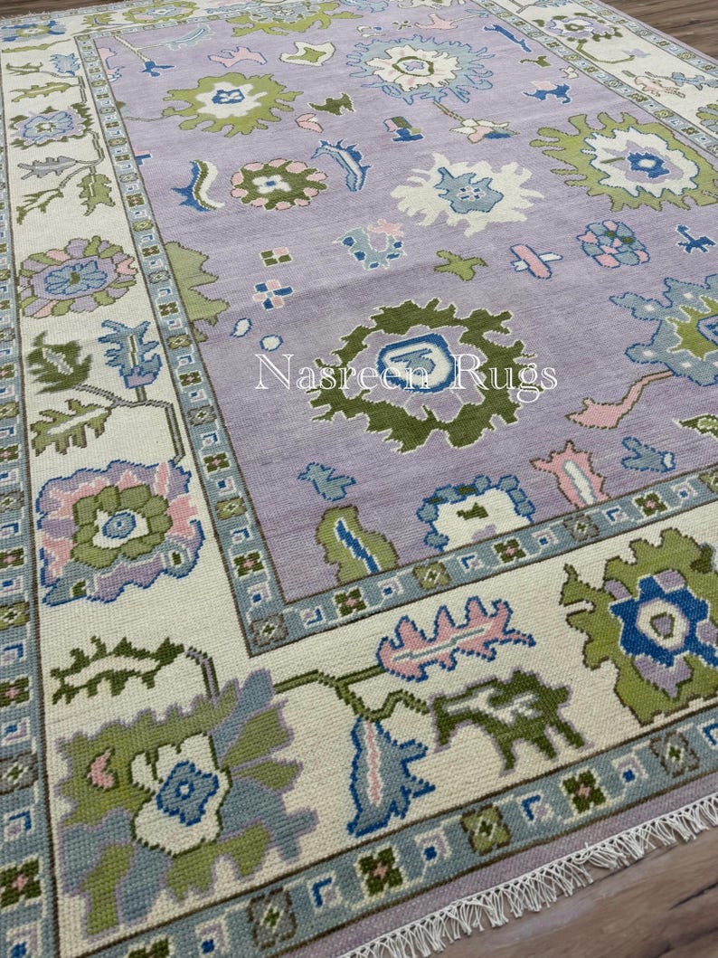 May include: A rectangular rug with a lavender central field and a cream border. The rug displays floral and geometric patterns in green, blue, and pink. The text "Nasreen Rugs" is visible on the rug.