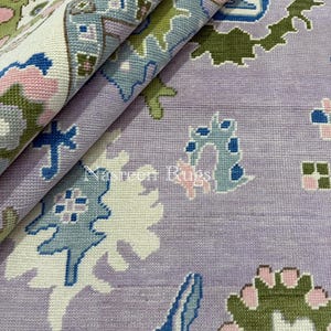 May include: A rolled-up, intricately patterned rug in shades of lavender, white, green, blue, and pink. The design features floral and geometric motifs. The text "Nasreen Rugs" is visible on the rug.