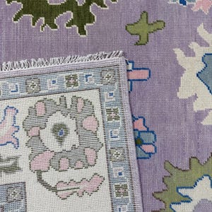 May include: A section of a patterned rug with a lavender background. The rug features floral and geometric designs in shades of green, blue, pink, and white. The edge of the rug is visible, showing a detailed border.