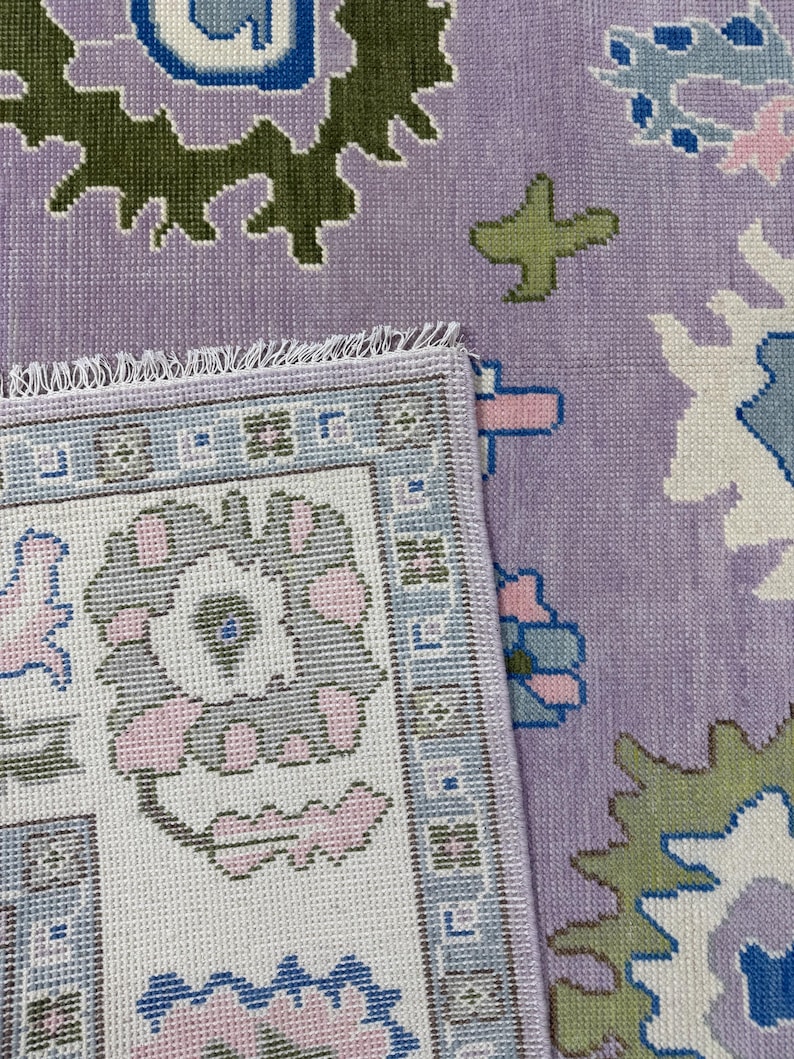 May include: A section of a patterned rug with a lavender background. The rug features floral and geometric designs in shades of green, blue, pink, and white. The edge of the rug is visible, showing a detailed border.