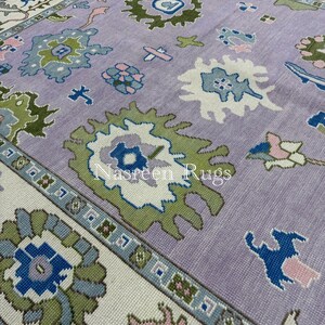 May include: A large, rectangular rug with a lavender central field and a cream border. The rug displays a repeating pattern of stylized floral and geometric shapes in green, blue, and pink. The text "Nasreen Rugs" is visible on the rug.