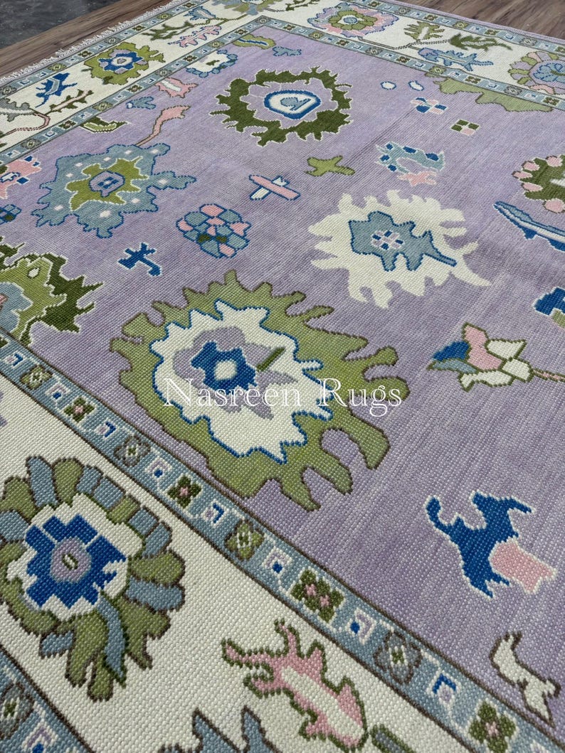 May include: A large, rectangular rug with a lavender central field and a cream border. The rug displays a repeating pattern of stylized floral and geometric shapes in green, blue, and pink. The text "Nasreen Rugs" is visible on the rug.