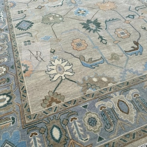 K&ouml;nnte beinhalten: Ein hellblauer und beiger Teppich mit einem Blumenmuster. Der Teppich hat einen Fransenrand und ist aus Wolle.