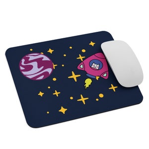 May include: A dark blue mouse pad with a pink rocket ship with a white cat inside, a purple planet, and yellow stars.