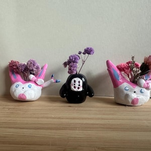 Puede incluir: Tres macetas de cerámica con forma de personajes de dibujos animados. Las macetas están pintadas de rosa, blanco y azul. Las macetas están llenas de flores secas.