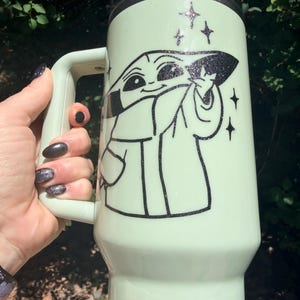 May include: A light green travel mug featuring a black and white illustration of a character from a popular science fiction series. The mug has a handle and a black lid. Glittery star accents surround the character's image.