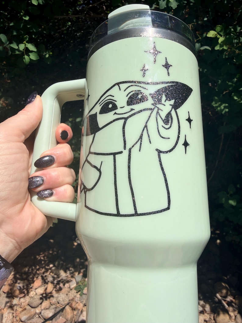 May include: A light green travel mug featuring a black and white illustration of a character from a popular science fiction series. The mug has a handle and a black lid. Glittery star accents surround the character's image.