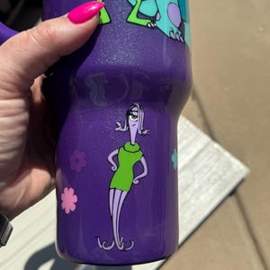 Puede incluir: Vaso t&eacute;rmico morado con los personajes de dibujos animados Mike Wazowski, Sulley y Roz de Monstruos S.A. El vaso tiene un acabado brillante y un asa. Tambi&eacute;n se ven detalles de flores rosas y azules.