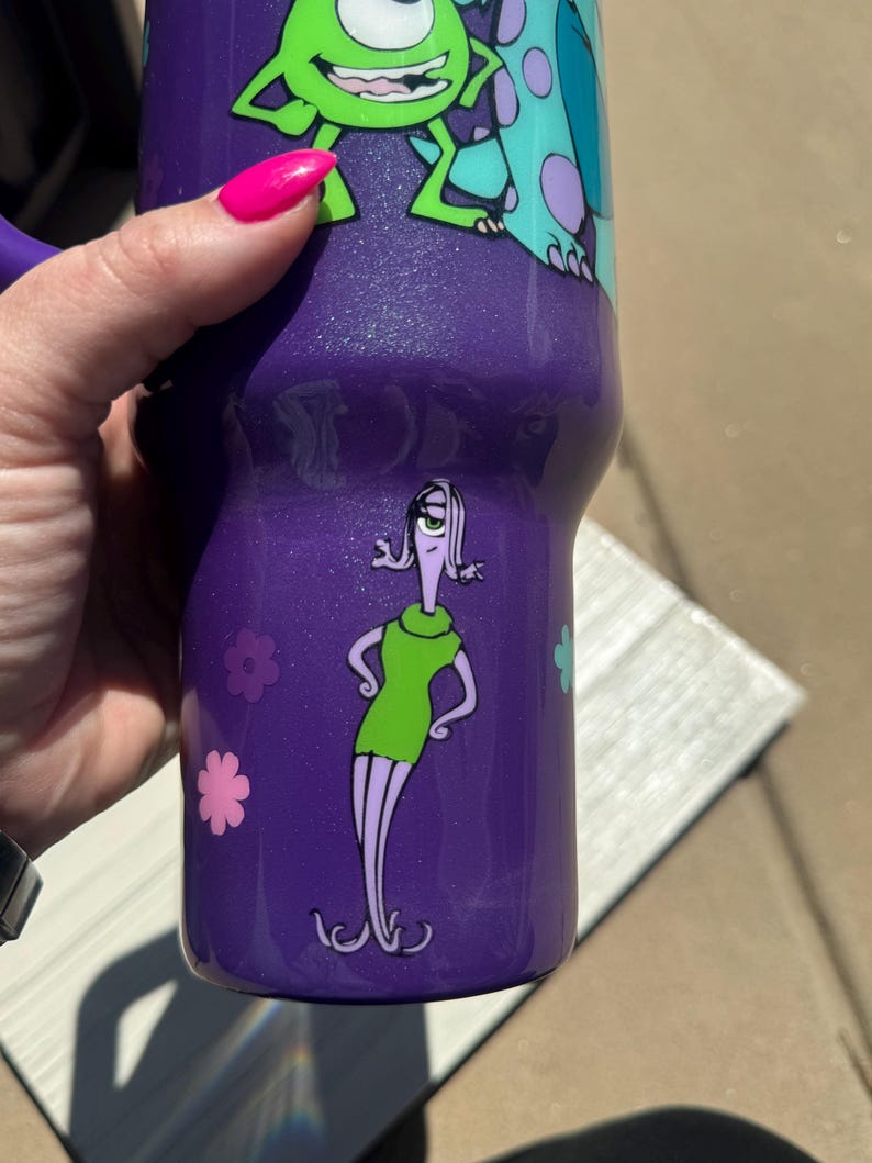 Puede incluir: Vaso t&eacute;rmico morado con los personajes de dibujos animados Mike Wazowski, Sulley y Roz de Monstruos S.A. El vaso tiene un acabado brillante y un asa. Tambi&eacute;n se ven detalles de flores rosas y azules.
