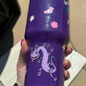 Puede incluir: Vaso morado con purpurina y calcoman&iacute;as de personajes de dibujos animados. El vaso presenta un personaje de dibujos animados de lagarto morado, calcoman&iacute;as de flores rosas y blancas y otras calcoman&iacute;as de dibujos animados. El vaso es cil&iacute;ndrico y parece estar hecho de acero inoxidable.