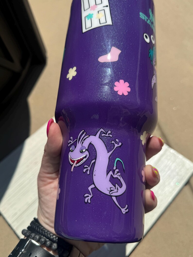 Puede incluir: Vaso morado con purpurina y calcoman&iacute;as de personajes de dibujos animados. El vaso presenta un personaje de dibujos animados de lagarto morado, calcoman&iacute;as de flores rosas y blancas y otras calcoman&iacute;as de dibujos animados. El vaso es cil&iacute;ndrico y parece estar hecho de acero inoxidable.