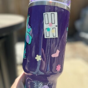 Puede incluir: Un vaso morado con dise&ntilde;os de personajes de dibujos animados. Tiene una tapa transparente y una pajita. El dise&ntilde;o incluye una puerta con acentos coloridos, flores, un calcet&iacute;n y un monstruo de dibujos animados.