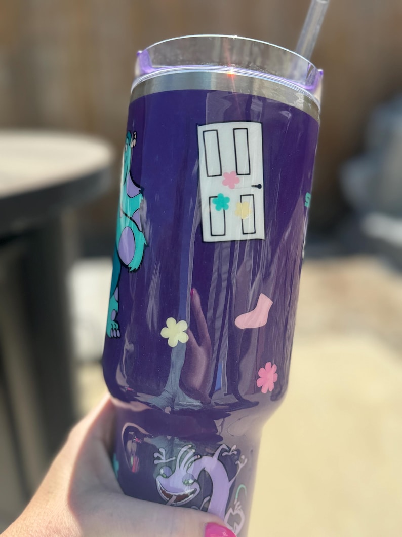 Puede incluir: Un vaso morado con dise&ntilde;os de personajes de dibujos animados. Tiene una tapa transparente y una pajita. El dise&ntilde;o incluye una puerta con acentos coloridos, flores, un calcet&iacute;n y un monstruo de dibujos animados.