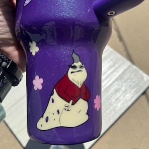 Puede incluir: Vaso morado con un dise&ntilde;o de personaje de dibujos animados. El vaso presenta un asa morada y un personaje de dibujos animados con gafas de sol, una chaqueta roja y un cuerpo blanco. Tambi&eacute;n hay dise&ntilde;os de flores rosas y blancas.