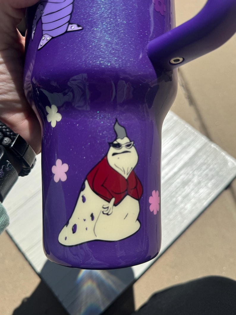 Puede incluir: Vaso morado con un dise&ntilde;o de personaje de dibujos animados. El vaso presenta un asa morada y un personaje de dibujos animados con gafas de sol, una chaqueta roja y un cuerpo blanco. Tambi&eacute;n hay dise&ntilde;os de flores rosas y blancas.