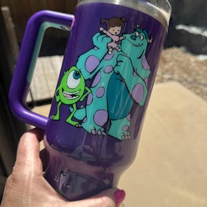 Puede incluir: Vaso t&eacute;rmico morado con asa, con los personajes de dibujos animados Mike Wazowski, Sulley y Boo de Monstruos S.A. El vaso tiene una tapa transparente y un asa morada con un acento turquesa.