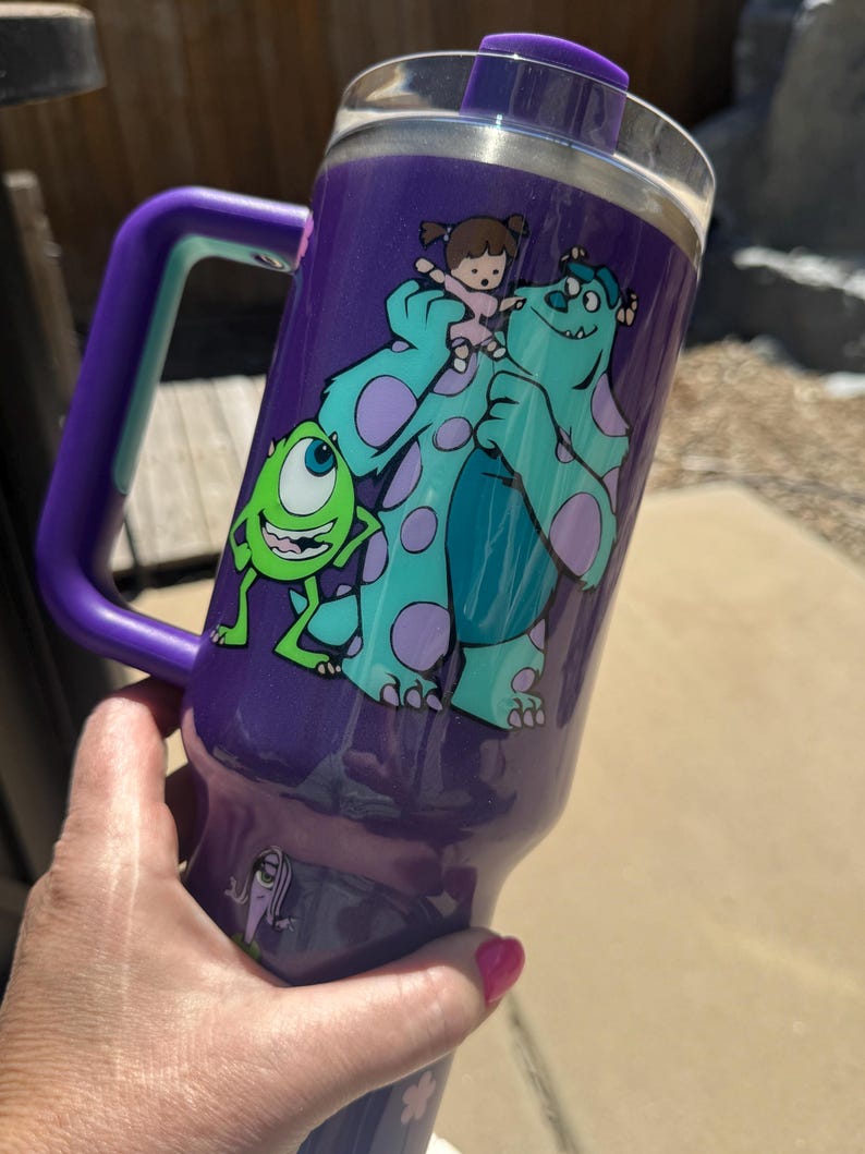 Puede incluir: Vaso t&eacute;rmico morado con asa, con los personajes de dibujos animados Mike Wazowski, Sulley y Boo de Monstruos S.A. El vaso tiene una tapa transparente y un asa morada con un acento turquesa.