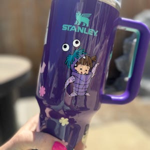 Puede incluir: Un vaso Stanley morado con asa y tapa. El vaso presenta un dise&ntilde;o de personaje de dibujos animados y el logotipo de Stanley. El asa y la tapa son moradas. El vaso tiene un borde de acero inoxidable.