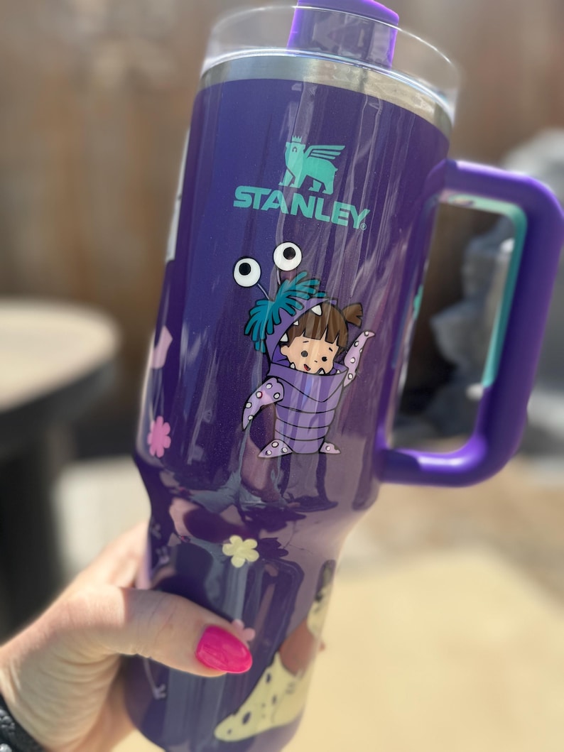 Puede incluir: Un vaso Stanley morado con asa y tapa. El vaso presenta un dise&ntilde;o de personaje de dibujos animados y el logotipo de Stanley. El asa y la tapa son moradas. El vaso tiene un borde de acero inoxidable.