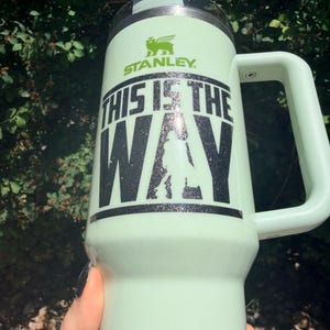 May include: A light green Stanley tumbler with a handle. The tumbler has the text "THIS IS THE WAY" in black glitter lettering, along with a silhouette and the Stanley logo. The tumbler is held by a person.