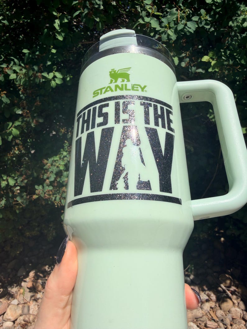May include: A light green Stanley tumbler with a handle. The tumbler has the text "THIS IS THE WAY" in black glitter lettering, along with a silhouette and the Stanley logo. The tumbler is held by a person.