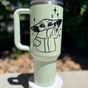May include: A light green travel mug with a black graphic of a character from a popular science fiction series. The mug has a handle, a clear lid, and a white base. The character is surrounded by black star graphics.