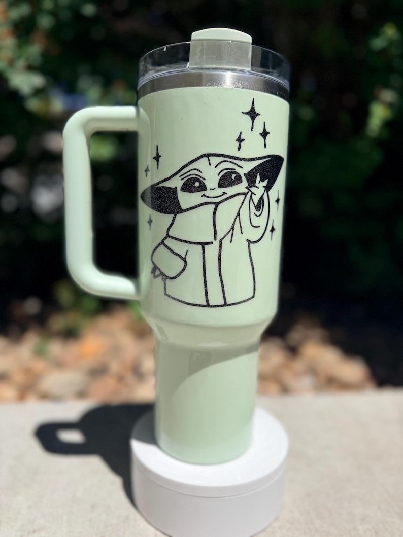 May include: A light green travel mug with a black graphic of a character from a popular science fiction series. The mug has a handle, a clear lid, and a white base. The character is surrounded by black star graphics.