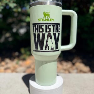 May include: A light green Stanley tumbler with a handle and lid. The tumbler features the text "THIS IS THE WAY" in black, along with a silhouette of a figure. The Stanley logo is visible above the text.