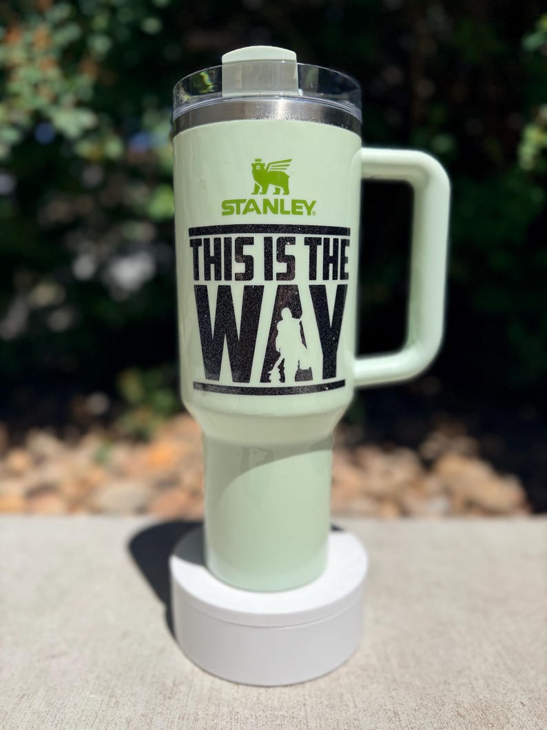 May include: A light green Stanley tumbler with a handle and lid. The tumbler features the text "THIS IS THE WAY" in black, along with a silhouette of a figure. The Stanley logo is visible above the text.